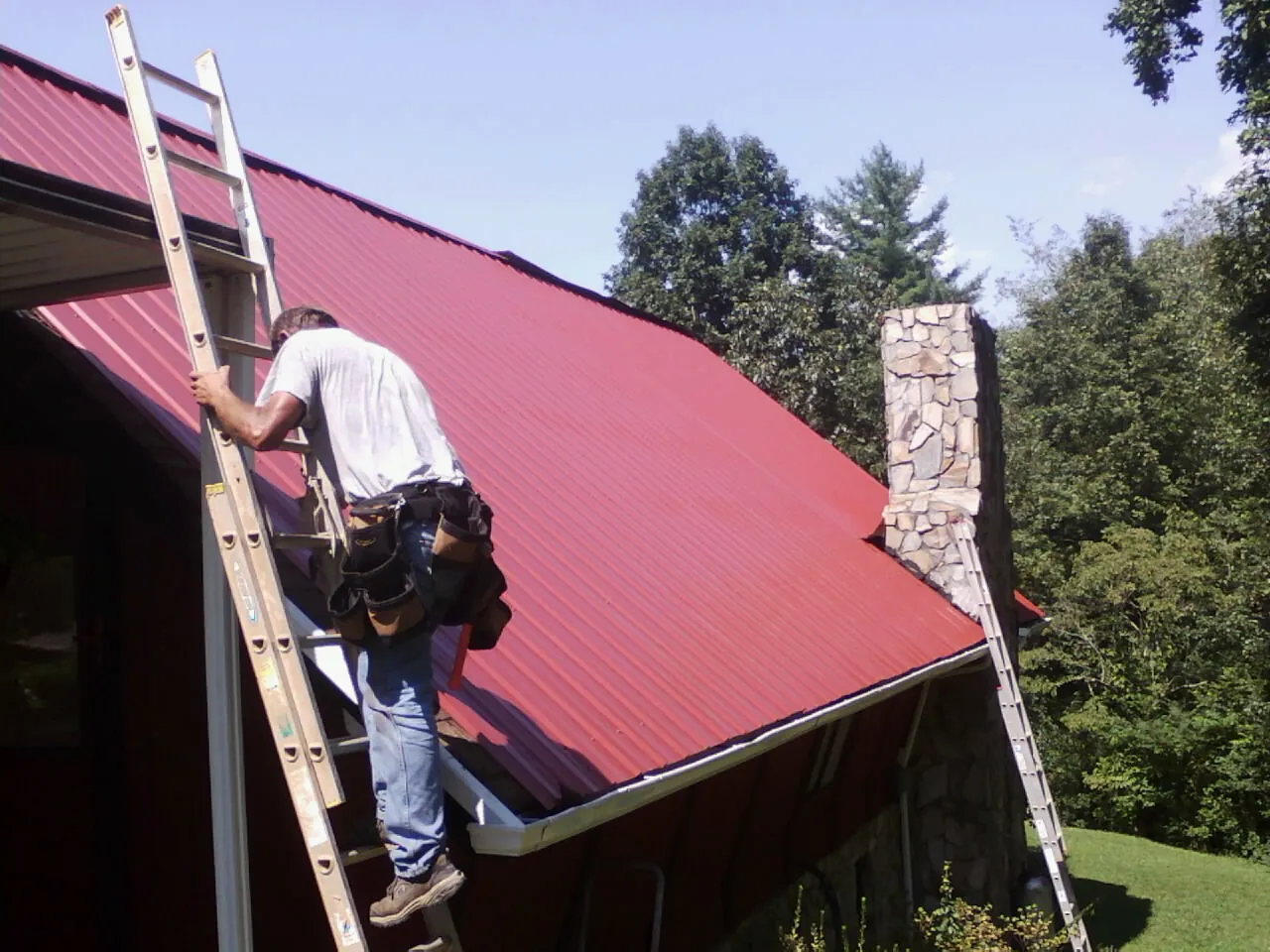 Roofer on a ladder beside a red metal roof and stone chimney — Soffit & Fascia Repair by Roofing Brothers in Woodland